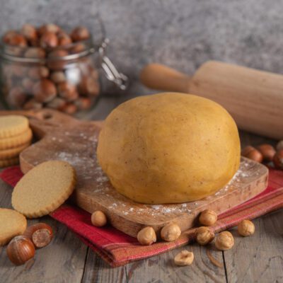 Pasta frolla alle nocciole formata a palla, con nocciole sparse e biscotti già cotti accanto