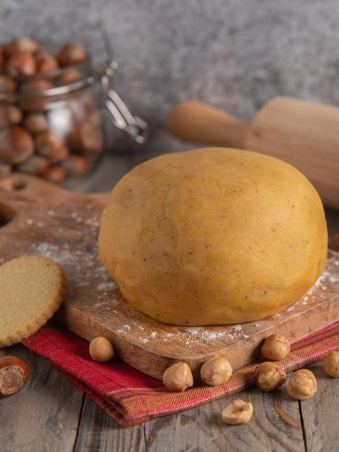 Pasta frolla alle nocciole formata a palla, con nocciole sparse e biscotti già cotti accanto