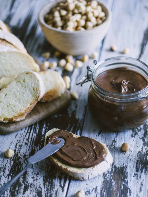 Crema spalmabile alle nocciole nel vasetto e spalmata su fetta di pane con nocciole intere