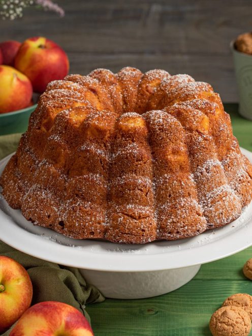 Ciambella con pesche e amaretti spolverata di zucchero a velo, con pesche fresche e biscotti intorno