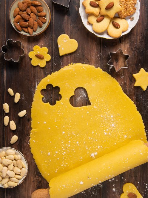 Pasta frolla alle mandorle stesa e tagliata con formine a cuore, fiore e stella, con biscotti pronti a lato