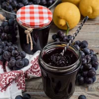 Confettura di uva fragola in vasetto aperto con cucchiaino, accanto a grappoli d'uva e limoni