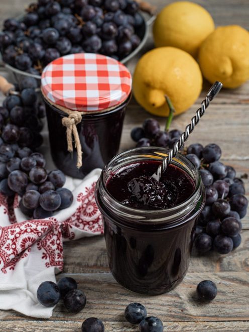 Confettura di uva fragola in vasetto aperto con cucchiaino, accanto a grappoli d'uva e limoni