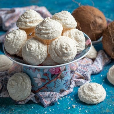 Biscotti al cocco dalla superficie granulosa e dorata, serviti in alzatina con noci di cocco sullo sfondo