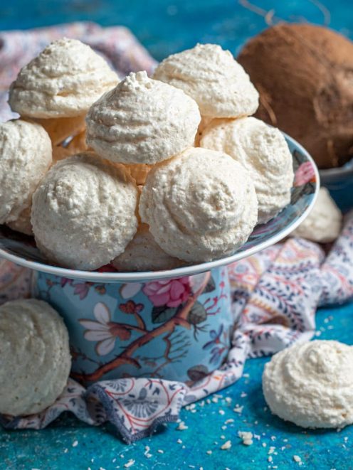 Biscotti al cocco dalla superficie granulosa e dorata, serviti in alzatina con noci di cocco sullo sfondo