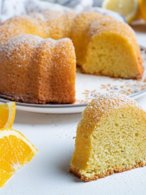 Dolce presto fatto a ciambella con zucchero a velo, una fetta in primo piano e spicchi d'arancia