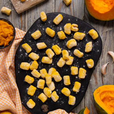 Gnocchi di zucca crudi rigati disposti su un tagliere infarinato, con zucca fresca e patate intorno