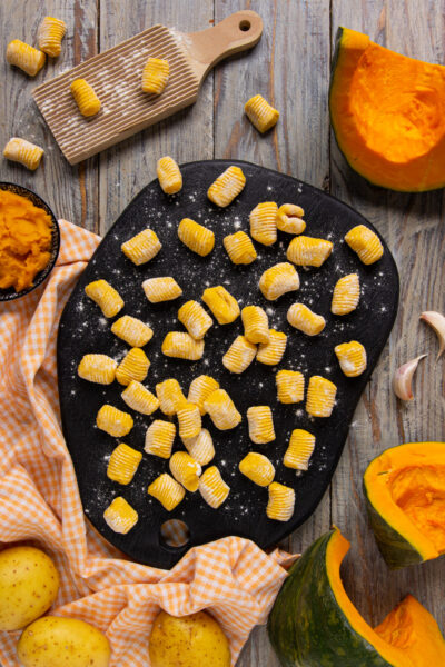 Gnocchi di zucca crudi rigati disposti su un tagliere infarinato, con zucca fresca e patate intorno
