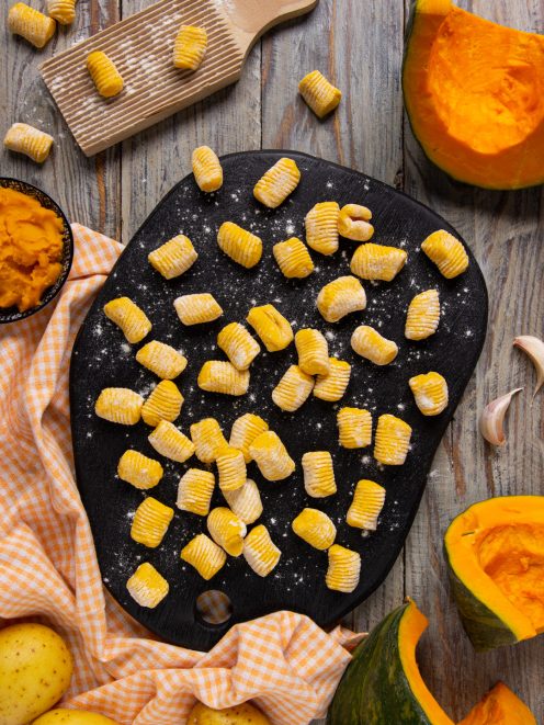 Gnocchi di zucca crudi rigati disposti su un tagliere infarinato, con zucca fresca e patate intorno