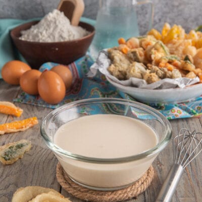 Pastella per fritti in una ciotola di vetro, circondata da verdure pastellate e anelli di cipolla fritti