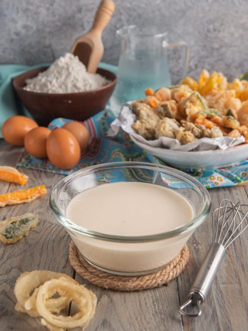 Pastella per fritti in una ciotola di vetro, circondata da verdure pastellate e anelli di cipolla fritti
