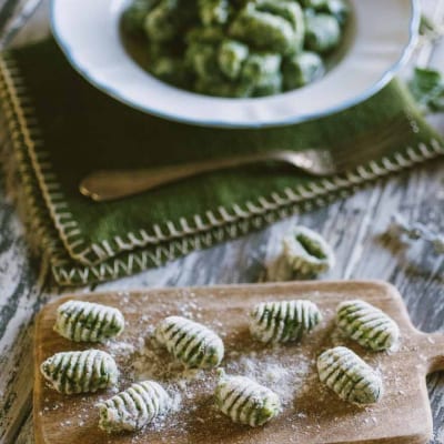 Gnocchi di spinaci rigati su tagliere infarinato, con piatto di gnocchi conditi sullo sfondo