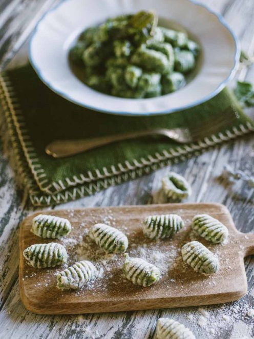 Gnocchi di spinaci rigati su tagliere infarinato, con piatto di gnocchi conditi sullo sfondo