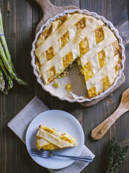 Quiche alle verdure con copertura a griglia dorata, una fetta servita a parte con asparagi freschi