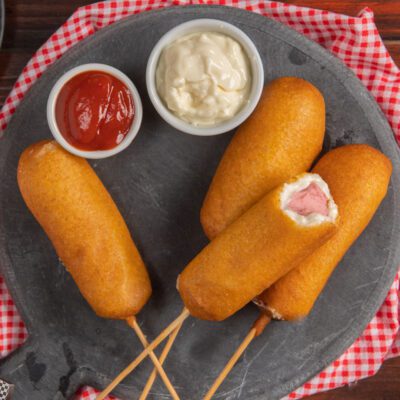 Corn dog fritti su stecco con wurstel visibile all'interno, serviti con ketchup e maionese