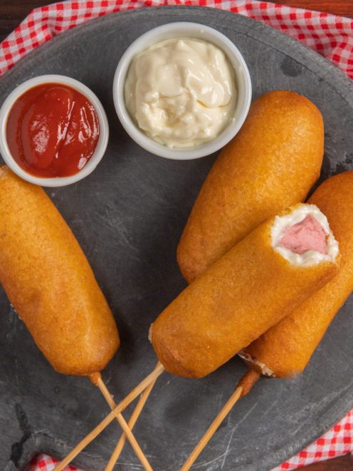 Corn dog fritti su stecco con wurstel visibile all'interno, serviti con ketchup e maionese