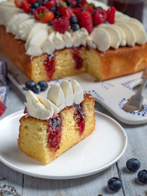 Poke cake farcita con confettura di frutti rossi, panna e fragole fresche in cima