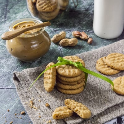 Biscotti al burro di arachidi impilati con fiocco verde, uno spezzato, accanto al vasetto di crema e arachidi sgusciate