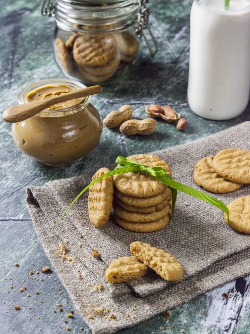 Biscotti al burro di arachidi impilati con fiocco verde, uno spezzato, accanto al vasetto di crema e arachidi sgusciate