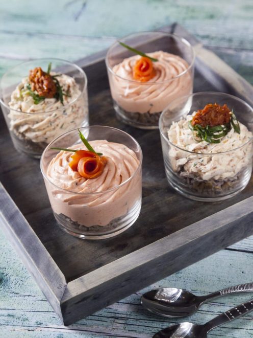 Mousse al salmone e mousse ai pomodori secchi servite in bicchieri, guarnite con salmone arrotolato ed erba cipollina