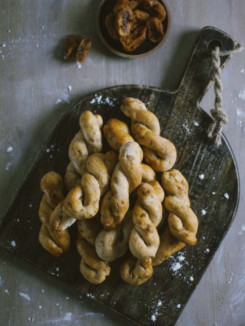 Treccine con fichi e noci impilate su tagliere, con fichi secchi sullo sfondo