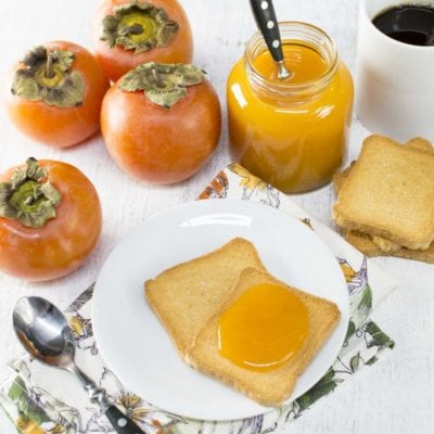 Composta di cachi spalmata su fette biscottate, con il vasetto aperto e i frutti freschi accanto