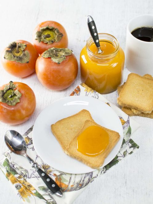 Composta di cachi spalmata su fette biscottate, con il vasetto aperto e i frutti freschi accanto