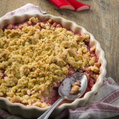 Crumble di rabarbaro con copertura dorata e sbriciolata, servito con un cucchiaio affondato nel ripieno rosa