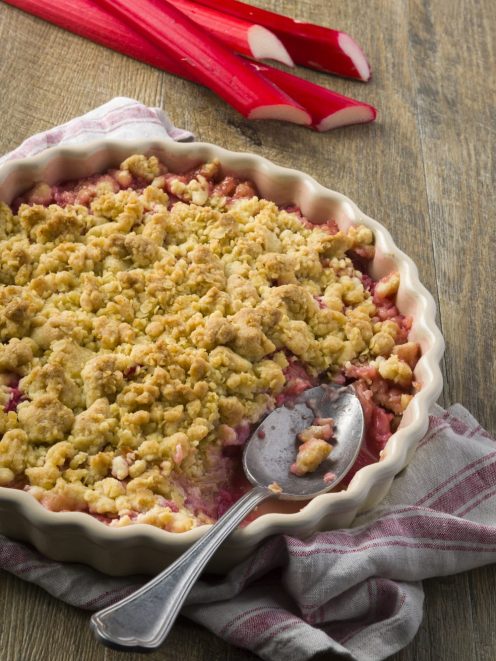 Crumble di rabarbaro con copertura dorata e sbriciolata, servito con un cucchiaio affondato nel ripieno rosa
