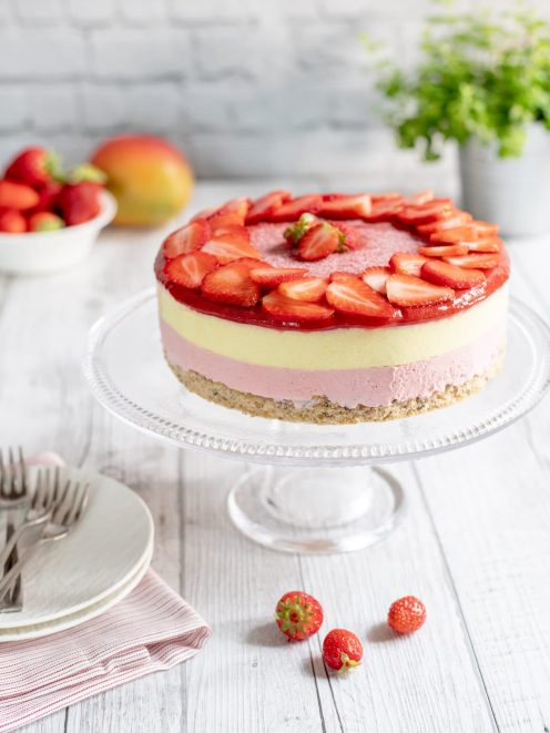 Mousse cake a strati con base di biscotto, mousse rosa e alla vaniglia, coronata da fragole fresche a fette