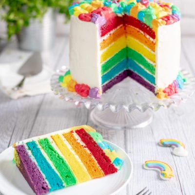 Rainbow cake a strati con glassa bianca e una fetta servita che rivela i sei strati colorati
