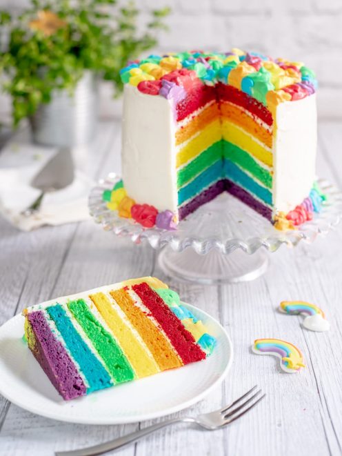 Rainbow cake a strati con glassa bianca e una fetta servita che rivela i sei strati colorati