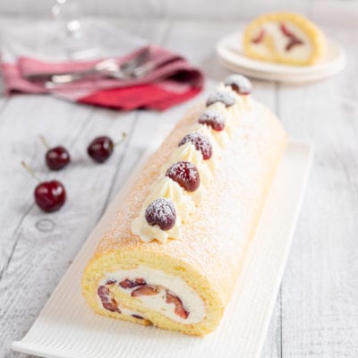 Rotolo alle ciliegie farcito con panna e frutta, decorato con ciuffi di crema e ciliegie spolverato di zucchero a velo