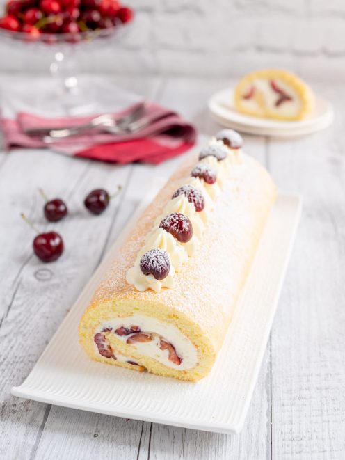 Rotolo alle ciliegie farcito con panna e frutta, decorato con ciuffi di crema e ciliegie spolverato di zucchero a velo
