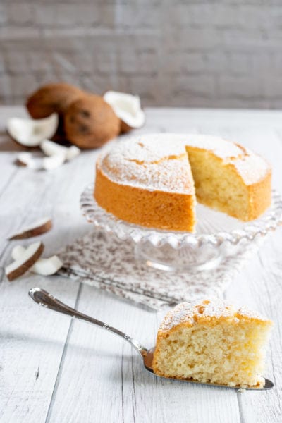 Torta al cocco spolverata di zucchero a velo, con una fetta servita in primo piano