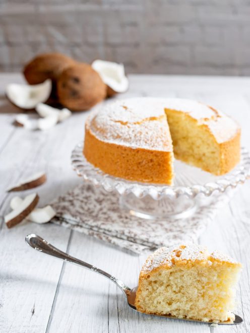 Torta al cocco spolverata di zucchero a velo, con una fetta servita in primo piano