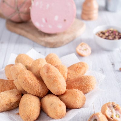 Crocchette di mortadella fritte e dorate, impilate su carta, con una spezzata che rivela il ripieno con pistacchi