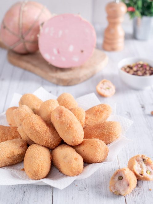 Crocchette di mortadella fritte e dorate, impilate su carta, con una spezzata che rivela il ripieno con pistacchi