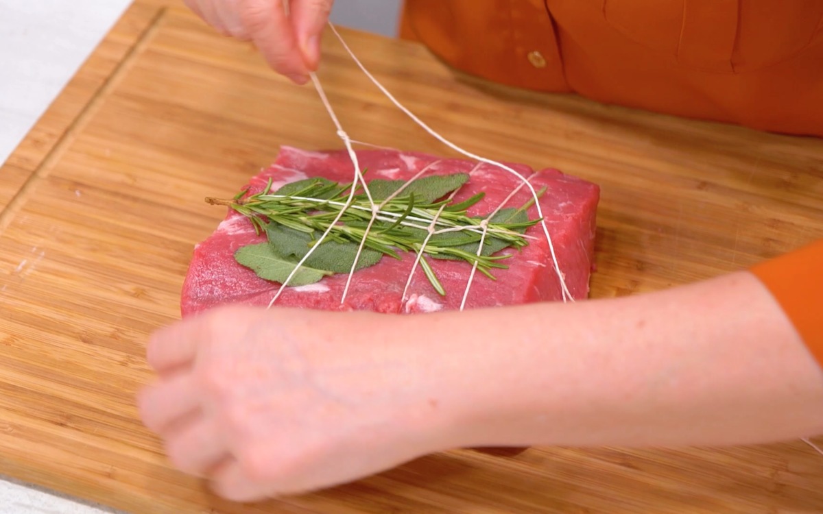 Mani che legano con spago un pezzo di carne cruda con rosmarino e salvia sopra