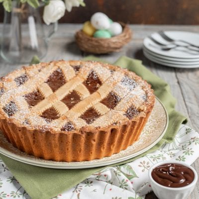 Pastiera al fondente con griglia di pasta frolla e zucchero a velo, accanto a una ciotolina di crema al cioccolato