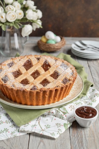 Pastiera al fondente con griglia di pasta frolla e zucchero a velo, accanto a una ciotolina di crema al cioccolato