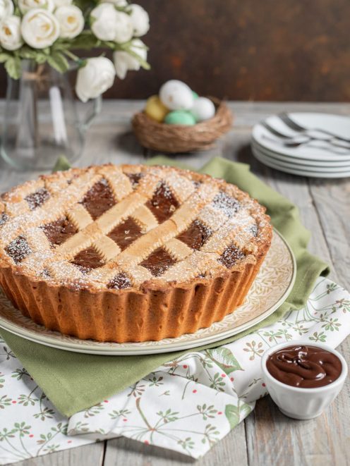 Pastiera al fondente con griglia di pasta frolla e zucchero a velo, accanto a una ciotolina di crema al cioccolato