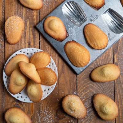 Madeleine dorate spolverate di zucchero a velo, servite su un piattino e sparse sul tavolo
