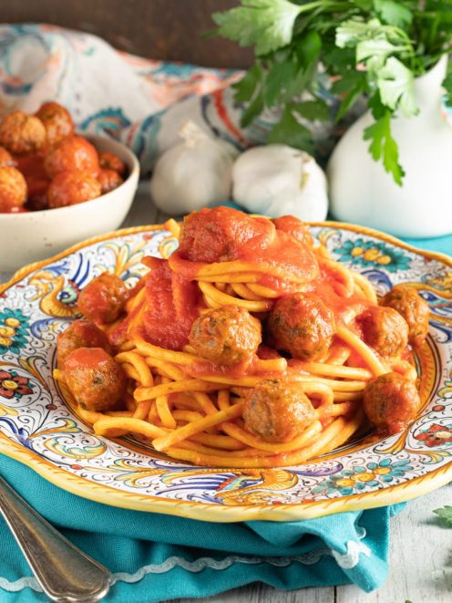 Spaghettoni al sugo con polpettine di alici disposti su piatto decorato accanto a una ciotola di polpettine