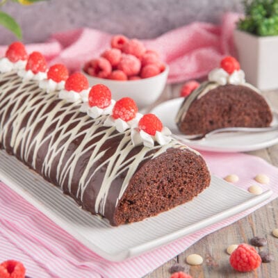 Tronchetto al cioccolato glassato con striature di cioccolato bianco e lamponi freschi su ciuffi di panna