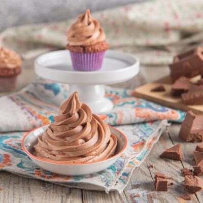 Ganache montata al cioccolato al latte in ciotolina, con cupcake glassati e cioccolato a pezzi sullo sfondo