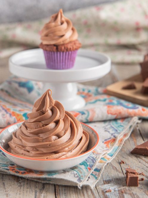 Ganache montata al cioccolato al latte in ciotolina, con cupcake glassati e cioccolato a pezzi sullo sfondo