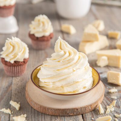 Ganache montata al cioccolato bianco in ciuffo a spirale, con pezzi di cioccolato e cupcake sullo sfondo
