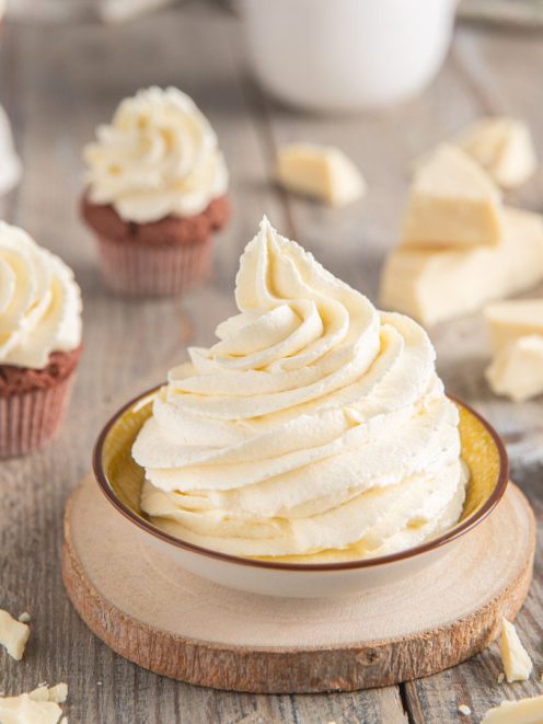 Ganache montata al cioccolato bianco in ciuffo a spirale, con pezzi di cioccolato e cupcake sullo sfondo