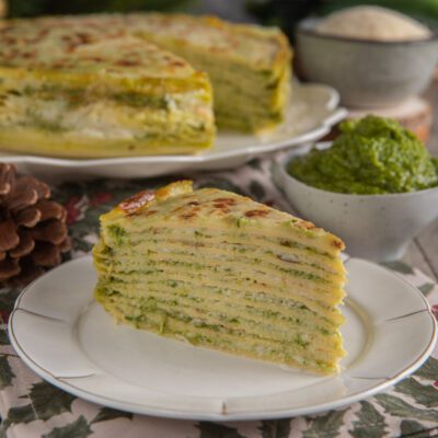 Torta di crespelle con pesto servita a fetta, con strati di verdure e superficie gratinata